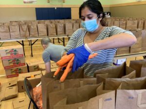 woman with a mask and gloves on begins to place a bundle of carrots in a brown grocery bag