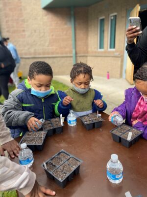 Children wearing masks and jackets, watering plants outside with small water bottles