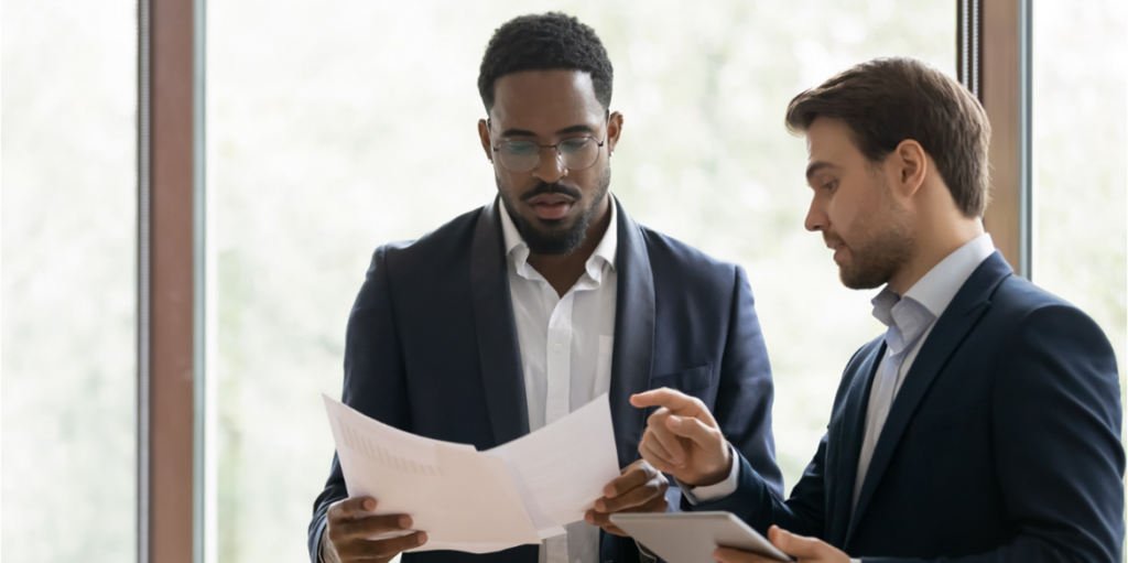 2 men in business suits standing in front of windows discussing something on the papers they are holding