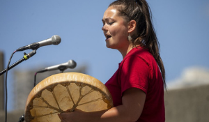 woman in a red outfit standing outside with a drum, singing into a microphone, with the blue sky above her